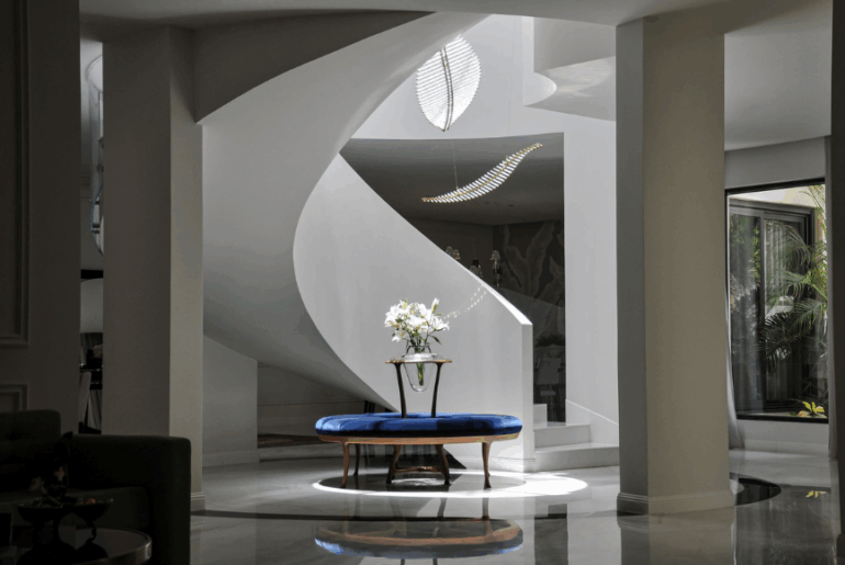 חלל כניסה יוקרתי עם גרם מדרגות ספירלי לבן, רצפת שיש מבריקה, ספסל מרופד כחול ושולחן מרכזי עם אגרטל פרחים תחת תאורה מעוצבת.