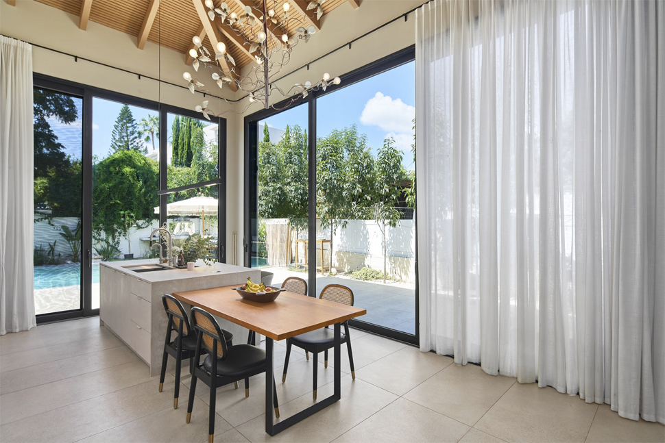 מטבח מודרני עם אי ומשטח בהיר, תקרה גבוהה עם קורות עץ וחלונות זכוכית גדולים הפונים לחצר עם בריכה