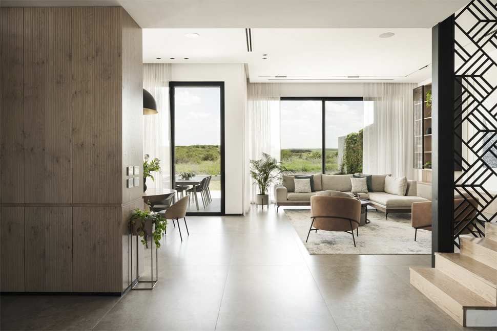 חלל מגורים מודרני עם סלון פתוח, מדרגות עץ ומפתחי זכוכית גדולים הפונים לחצר ירוקה